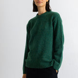 A woman wearing a dark green textured knit sweater and black trousers, standing against a plain white wall.
