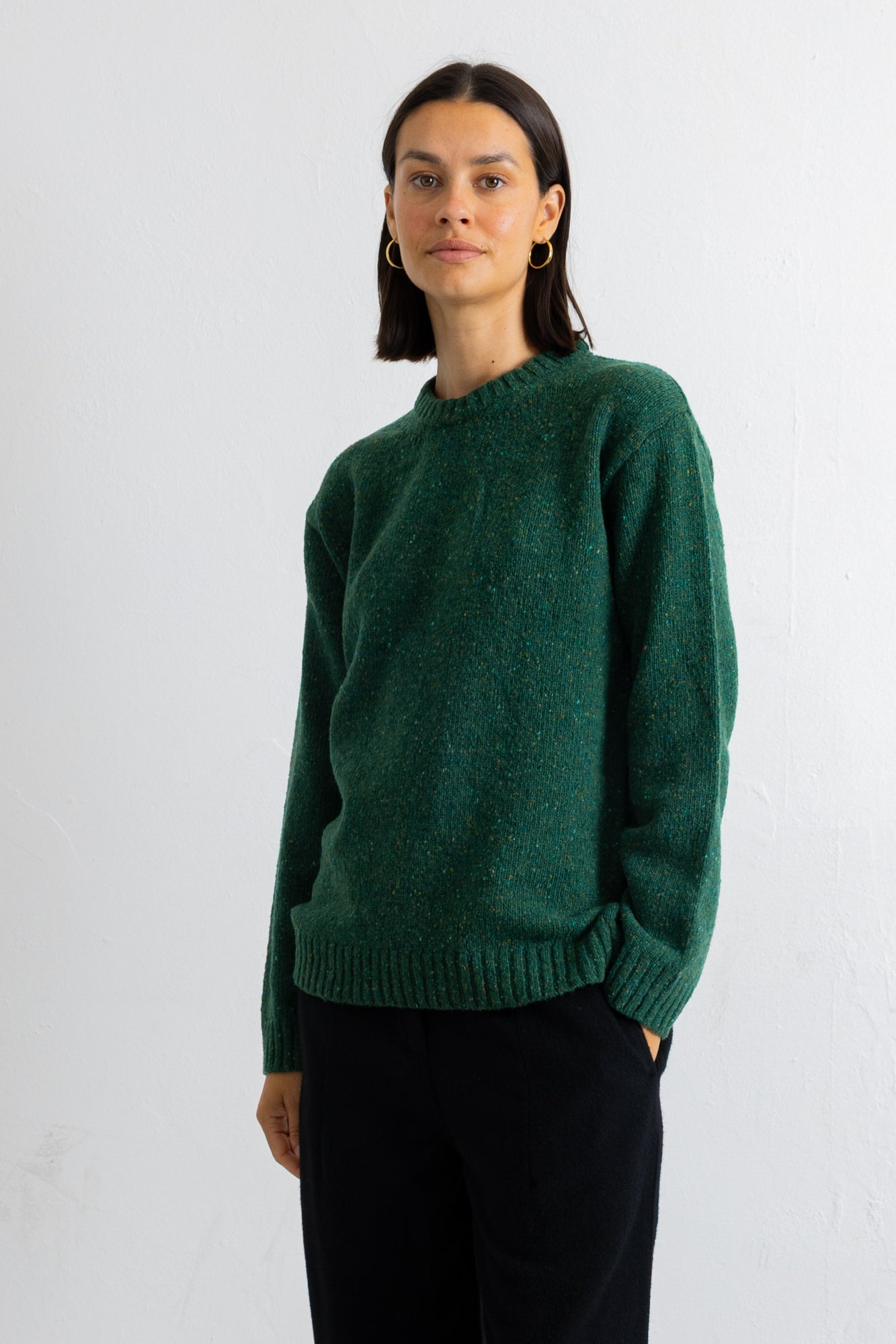 A woman wearing a dark green textured knit sweater and black trousers, standing against a plain white wall.