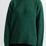 A person wearing a dark green heathered knit sweater with a crew neck and ribbed cuffs and hem, paired with black pants, against a plain white wall.