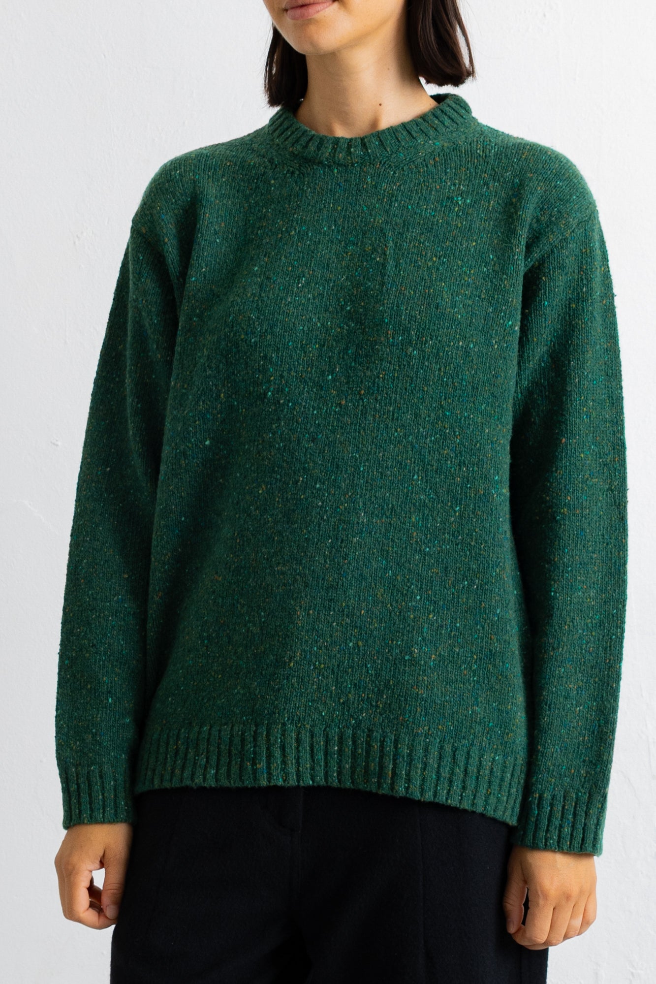 A person wearing a dark green heathered knit sweater with a crew neck and ribbed cuffs and hem, paired with black pants, against a plain white wall.
