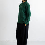 A woman stands in profile wearing a dark green knit sweater and loose black trousers against a plain white studio background.