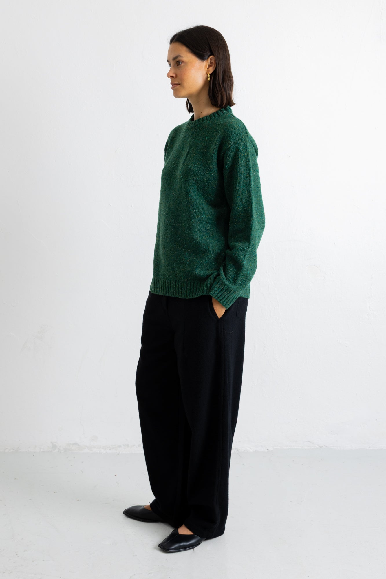 A woman stands in profile wearing a dark green knit sweater and loose black trousers against a plain white studio background.