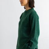 A woman wearing a dark green knit sweater and black pants, shown in profile against a plain white wall.