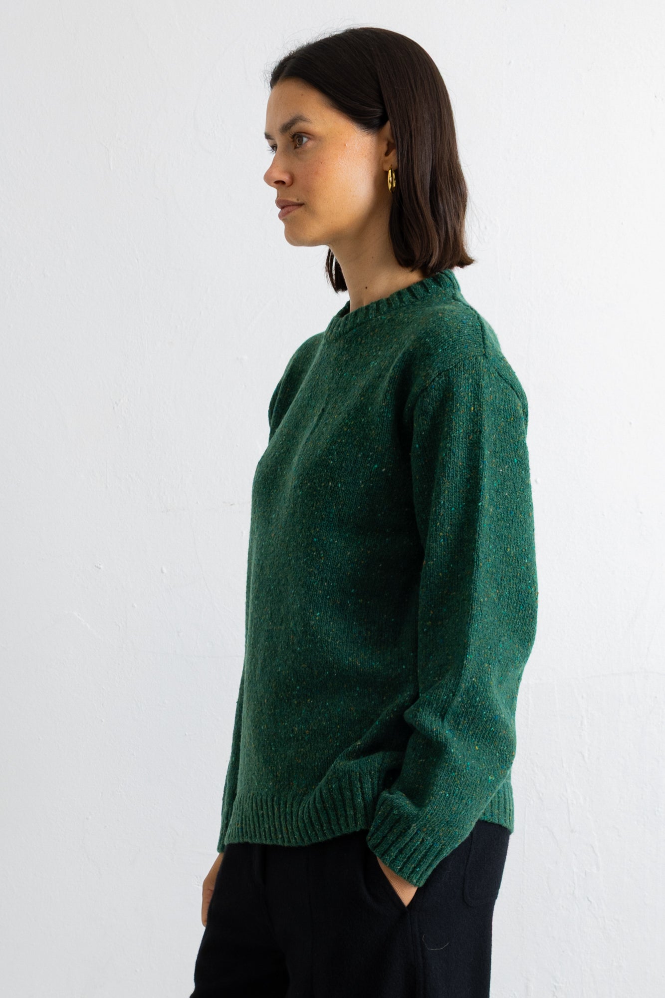 A woman wearing a dark green knit sweater and black pants, shown in profile against a plain white wall.