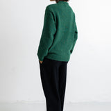 A woman stands in profile wearing a textured green knit sweater and black trousers against a plain white studio background.
