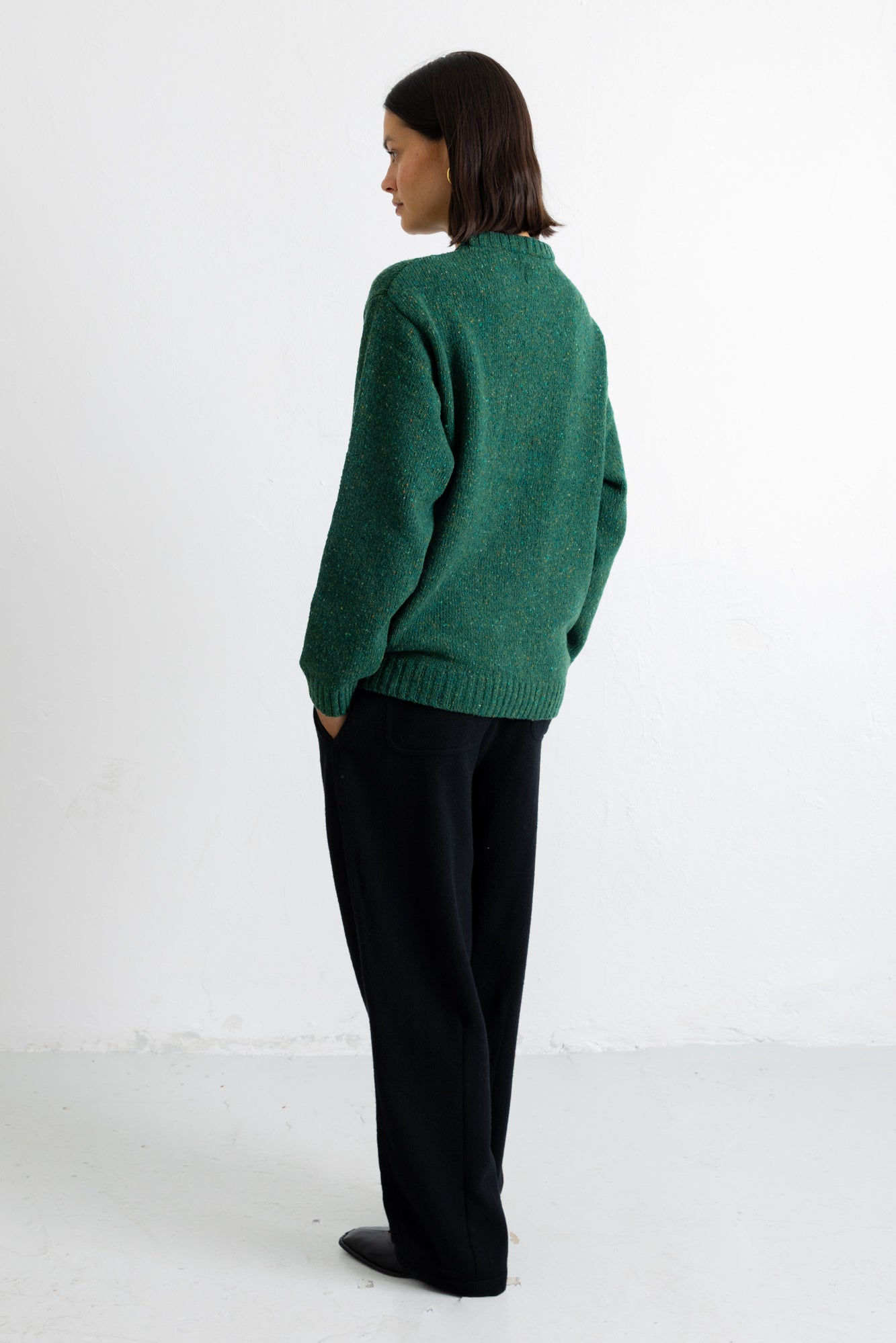 A woman stands in profile wearing a textured green knit sweater and black trousers against a plain white studio background.
