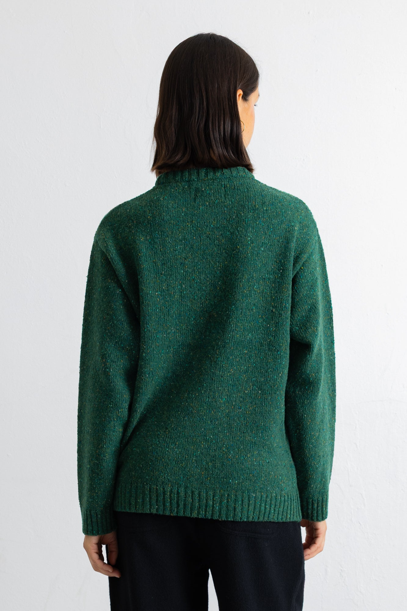 A person shown from the back wearing a dark green knit sweater and dark pants against a plain white studio background.