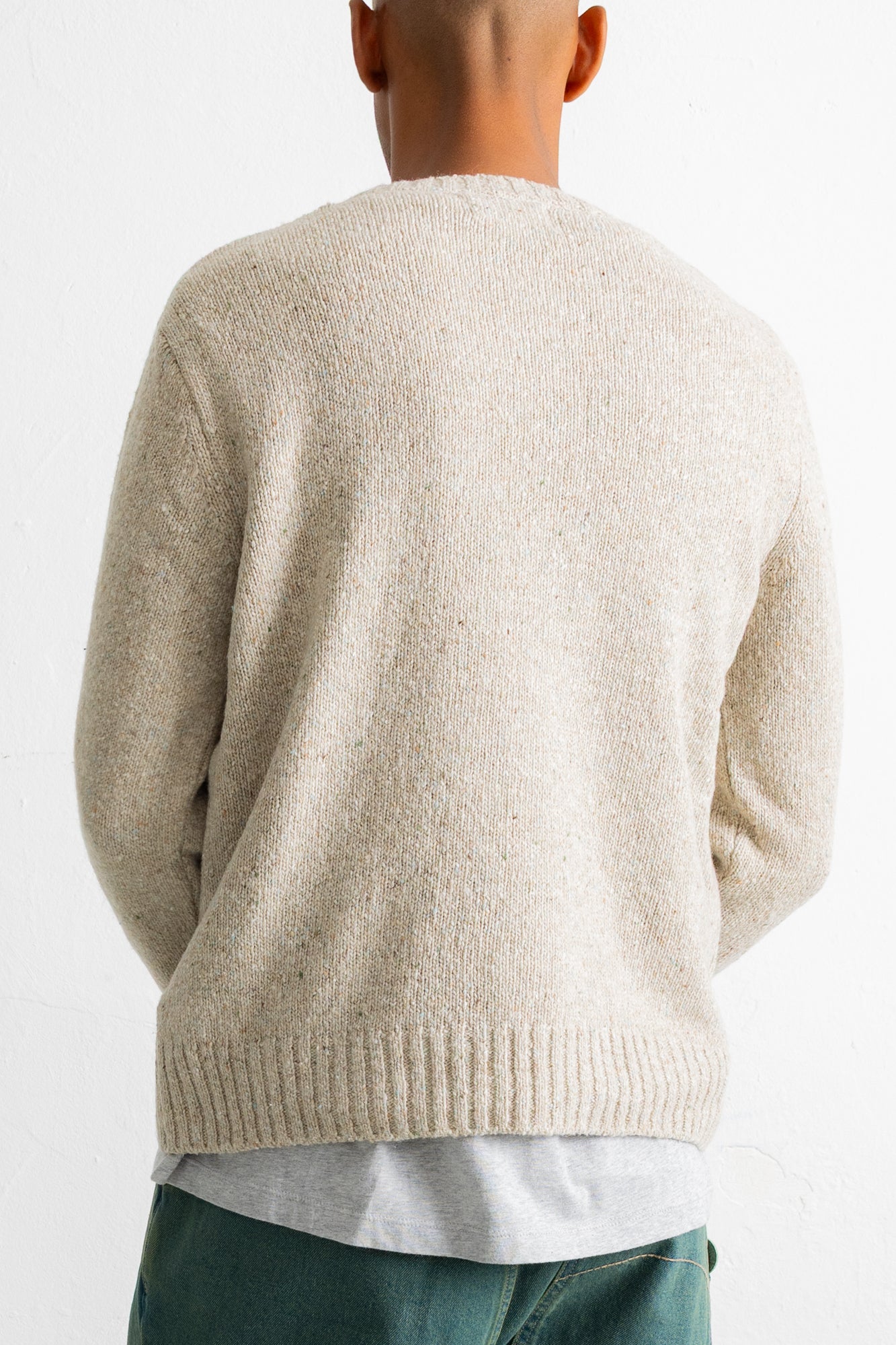 A person viewed from the back wearing a cream-colored knit sweater over a light gray shirt and teal pants, against a plain white background.