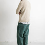 A man standing in profile wearing a light beige knit sweater, green trousers, and black-and-white sneakers against a plain white studio wall.