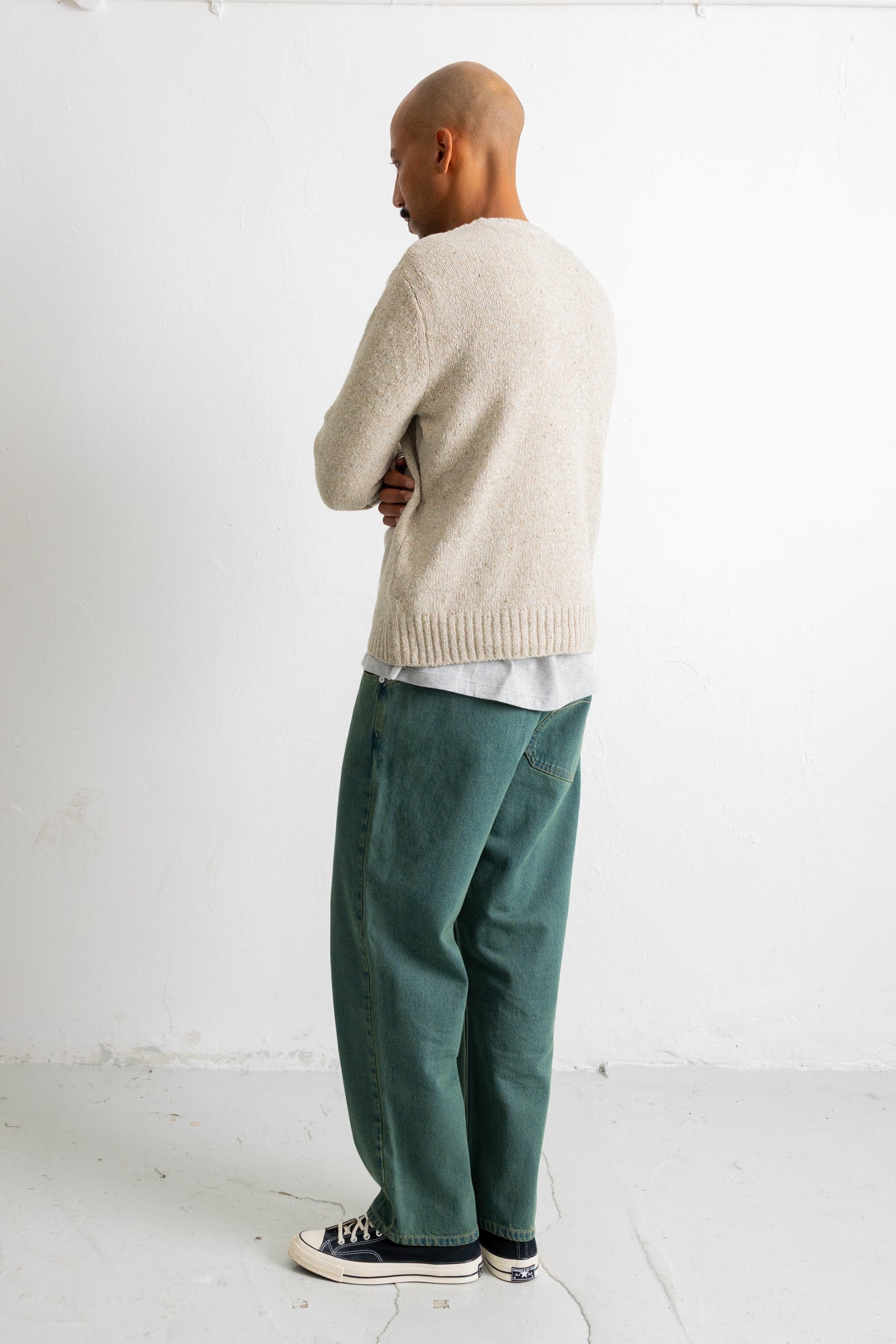 A man standing in profile wearing a light beige knit sweater, green trousers, and black-and-white sneakers against a plain white studio wall.