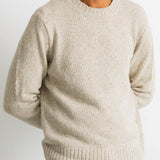 A man wearing a light beige crewneck knit sweater, paired with a light gray t-shirt and teal jeans, shown from the chest down against a plain white background.