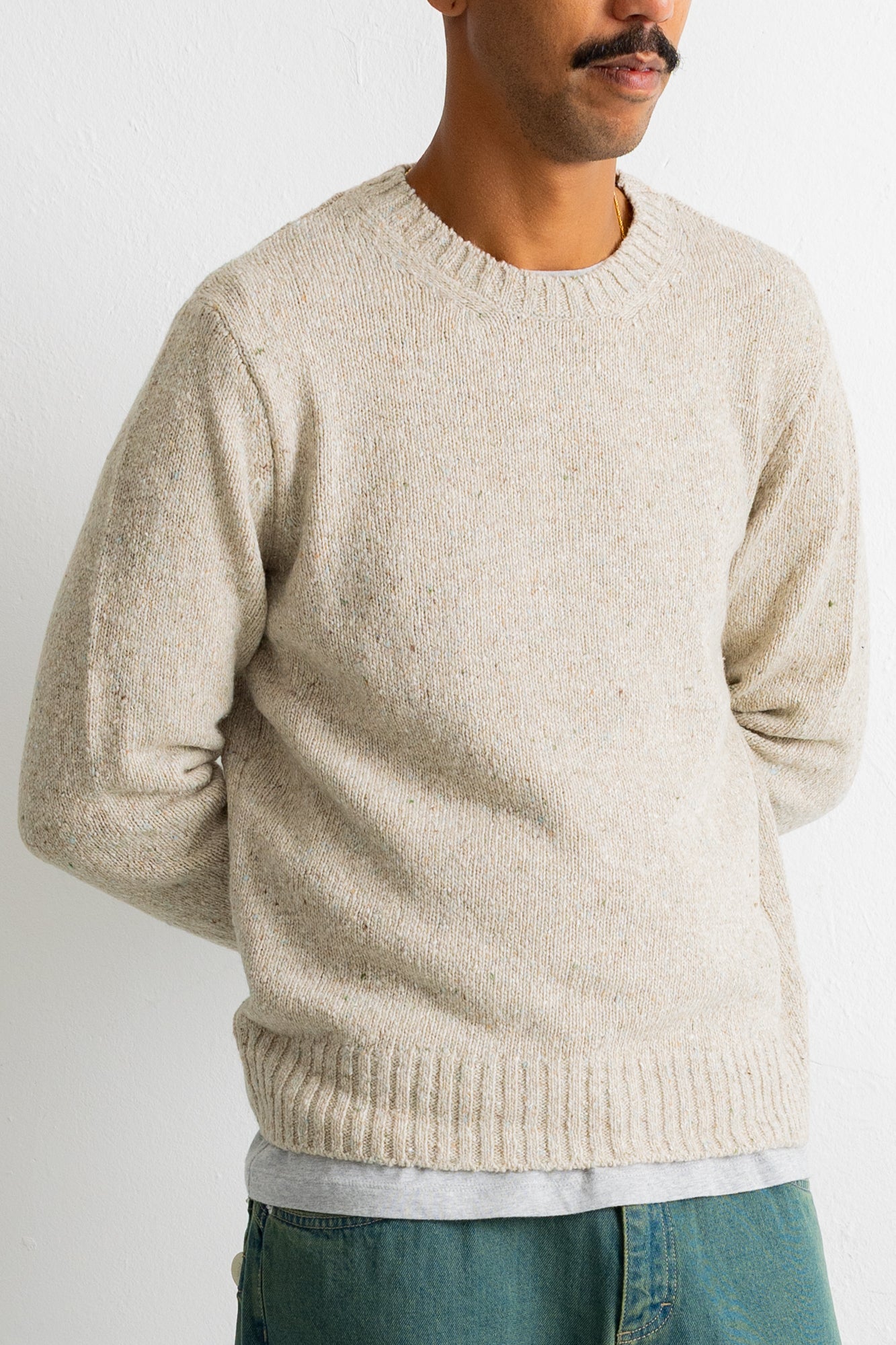 A man wearing a light beige crewneck knit sweater, paired with a light gray t-shirt and teal jeans, shown from the chest down against a plain white background.