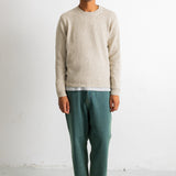 A man wearing a light beige knit crewneck sweater, green trousers, and black-and-white sneakers, standing against a plain white wall.