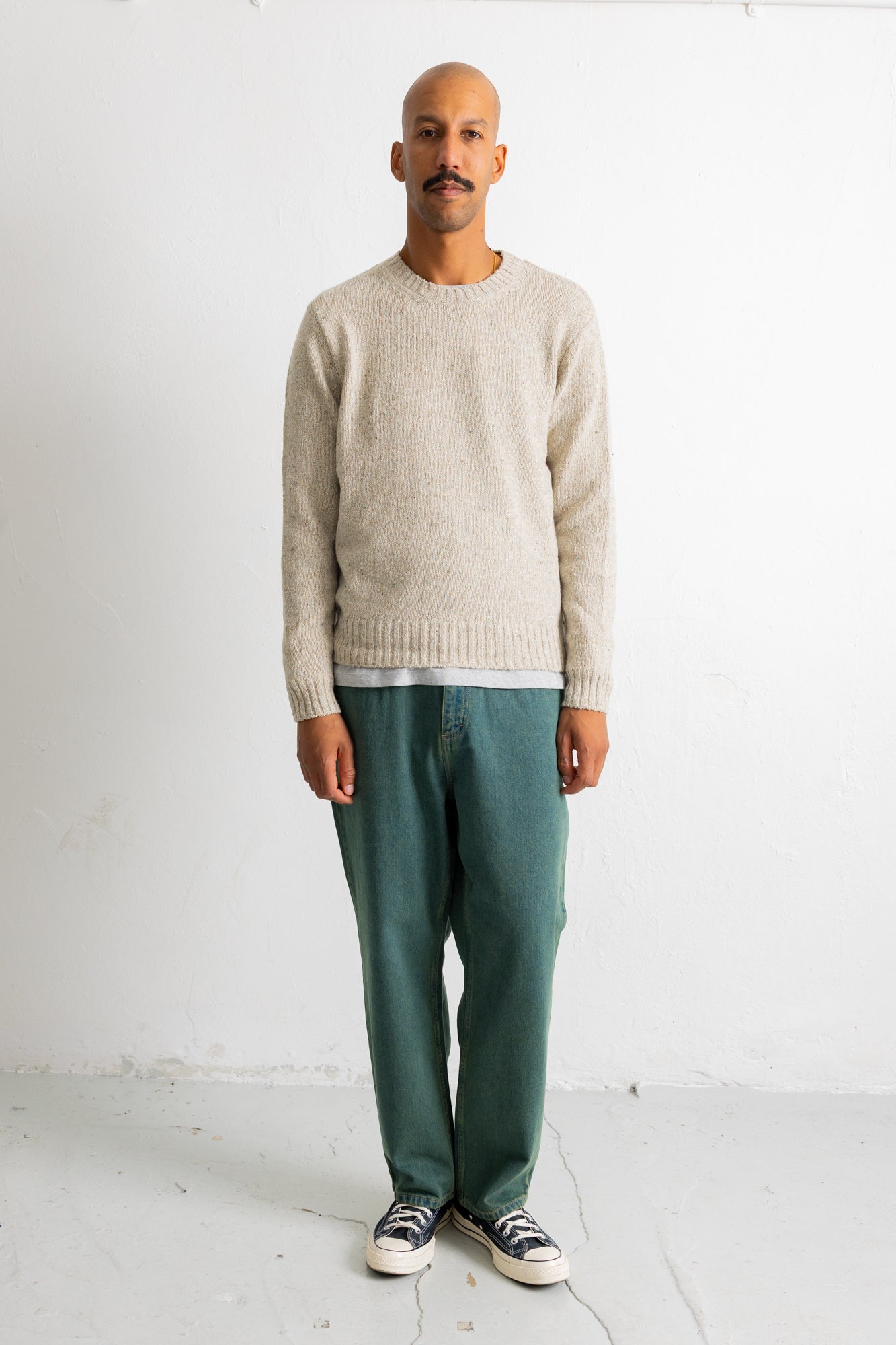 A man wearing a light beige knit crewneck sweater, green trousers, and black-and-white sneakers, standing against a plain white wall.
