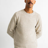 A man wearing a light beige crewneck knit sweater and teal pants, standing against a plain white wall.