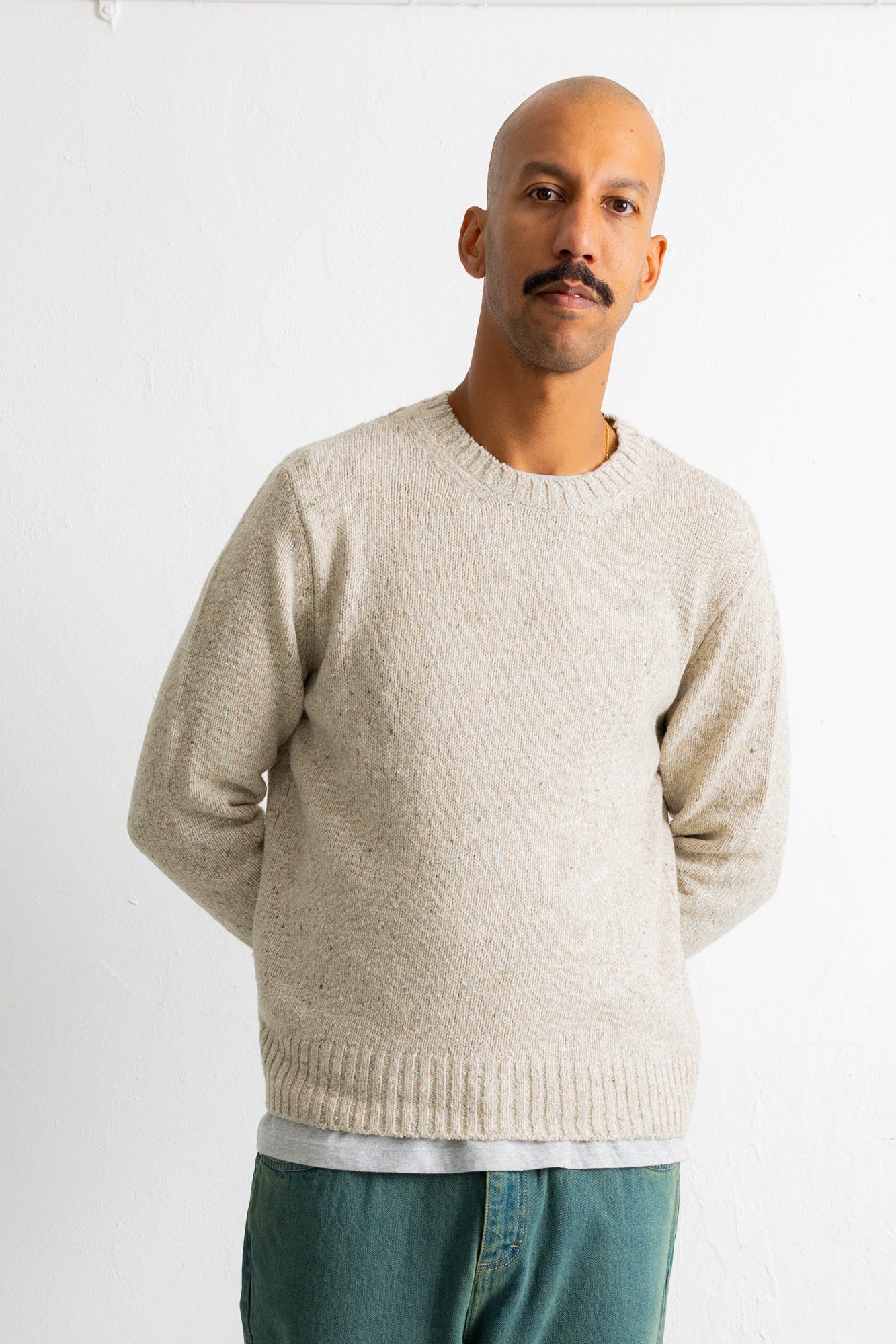 A man wearing a light beige crewneck knit sweater and teal pants, standing against a plain white wall.