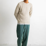A man wearing a light beige knit crewneck sweater, dark green wide-leg trousers, and black-and-white sneakers, standing against a plain white studio wall.