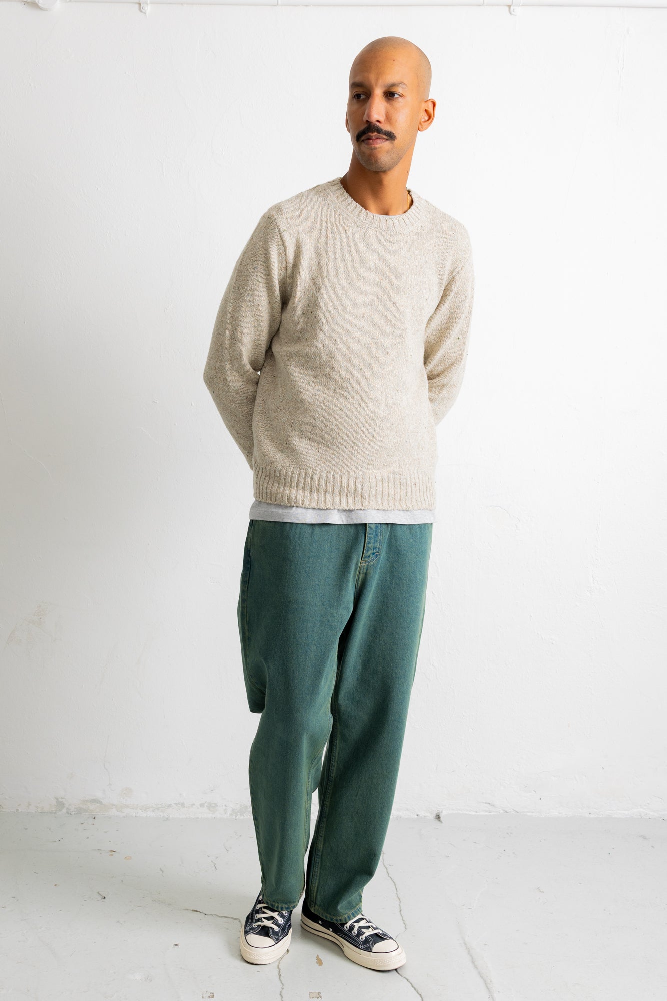 A man wearing a light beige knit crewneck sweater, dark green wide-leg trousers, and black-and-white sneakers, standing against a plain white studio wall.