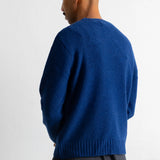 A man shown from the back wearing a vibrant blue knit sweater over a striped shirt and green pants, photographed against a plain white wall.