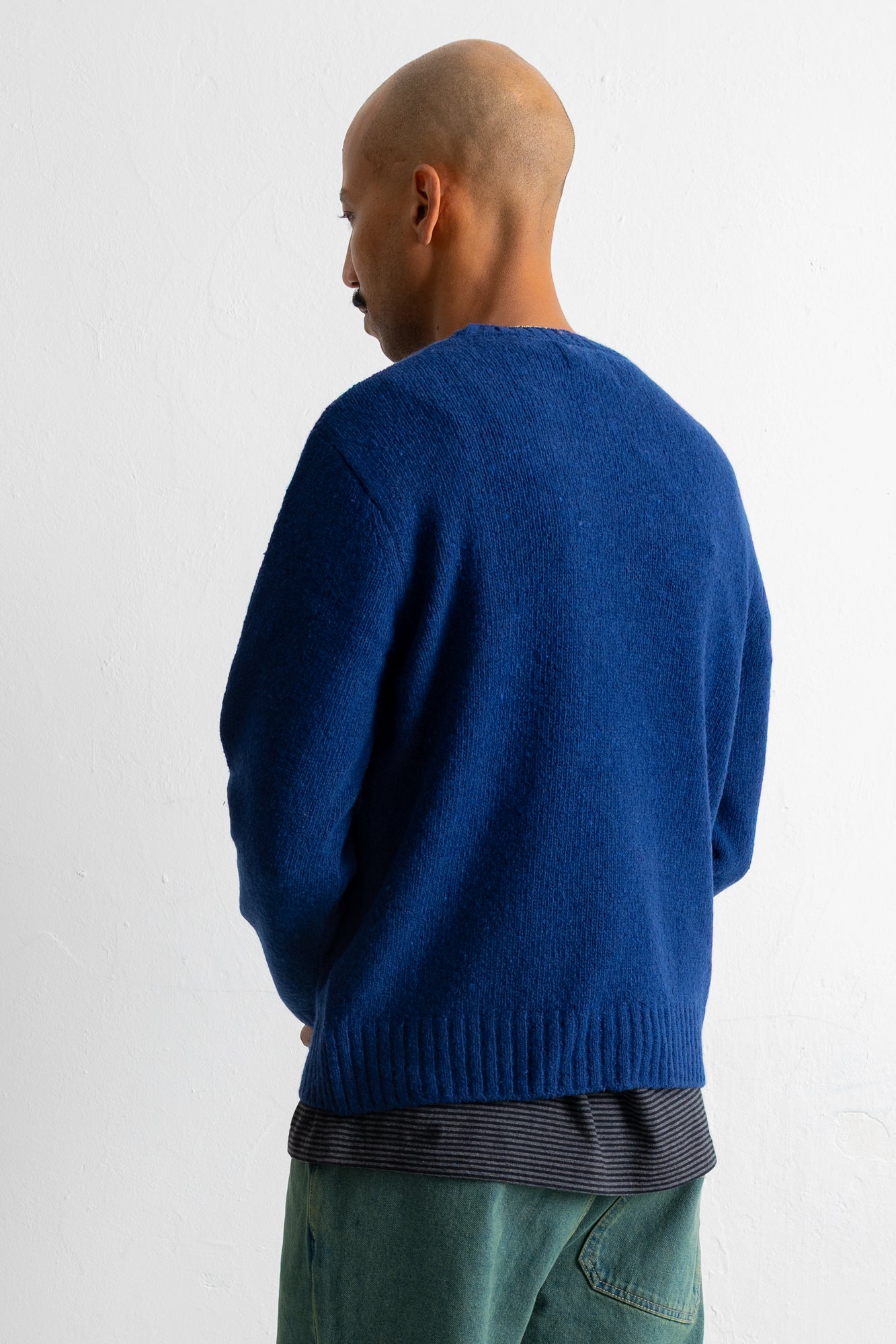 A man shown from the back wearing a vibrant blue knit sweater over a striped shirt and green pants, photographed against a plain white wall.