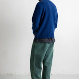 A man wearing a blue knit sweater, a dark gray t-shirt, green corduroy pants, and black canvas sneakers, standing in profile against a white studio wall.