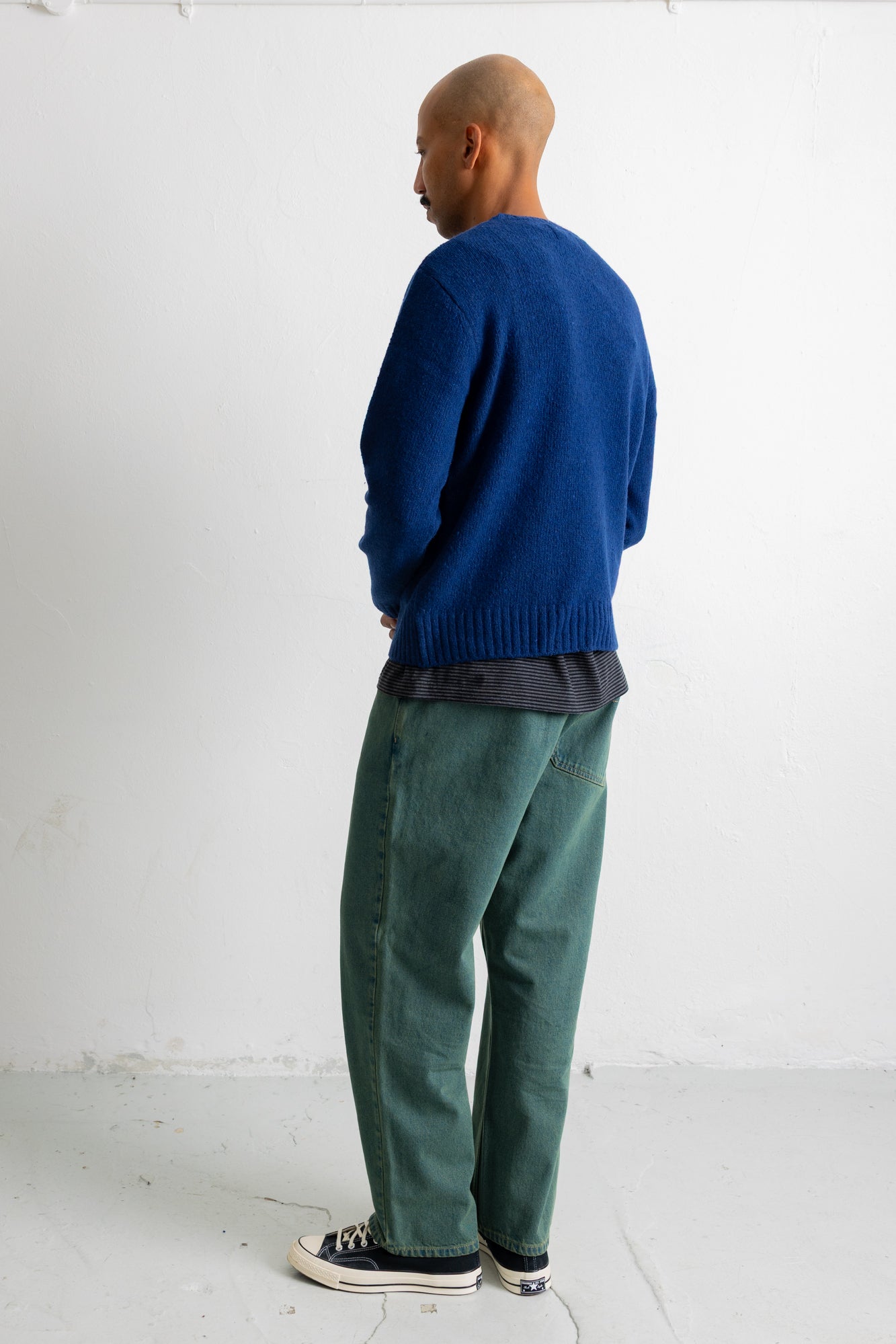 A man wearing a blue knit sweater, a dark gray t-shirt, green corduroy pants, and black canvas sneakers, standing in profile against a white studio wall.