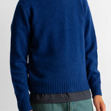 A man wearing a royal blue knit crewneck sweater, paired with green pants and a striped shirt hem showing at the waist, against a plain light background.