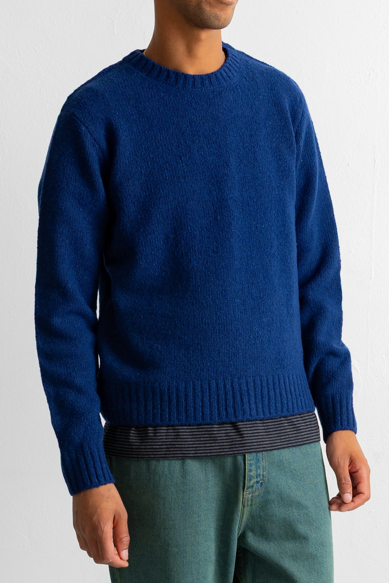 A man wearing a royal blue knit crewneck sweater, paired with green pants and a striped shirt hem showing at the waist, against a plain light background.