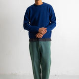 A man wearing a navy blue knit crewneck sweater, green trousers, and black-and-white sneakers, standing against a plain white studio wall.