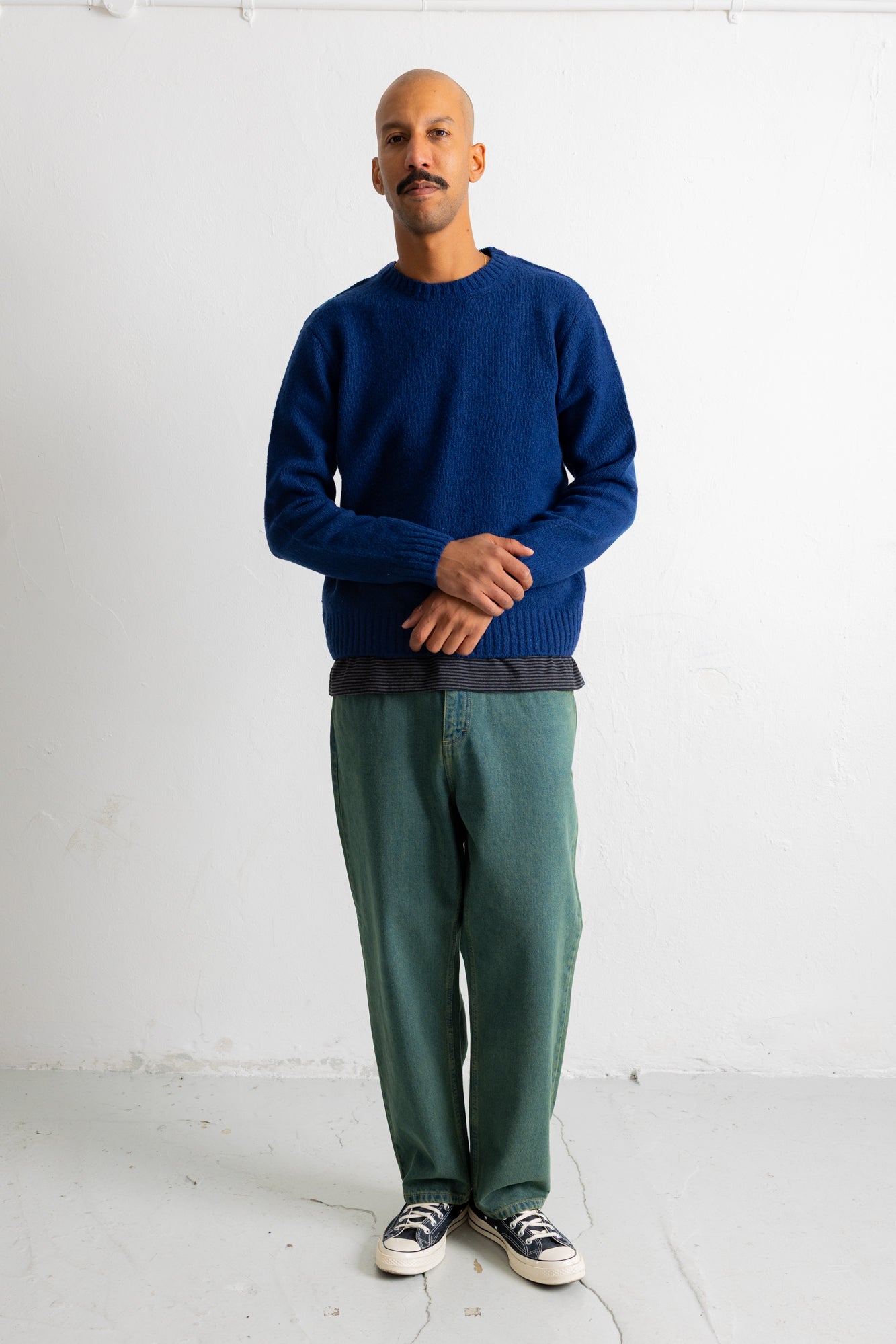 A man wearing a navy blue knit crewneck sweater, green trousers, and black-and-white sneakers, standing against a plain white studio wall.