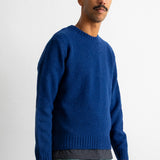 A man wearing a vibrant royal blue knit crewneck sweater, paired with green trousers and a striped undershirt, standing against a plain white studio background.