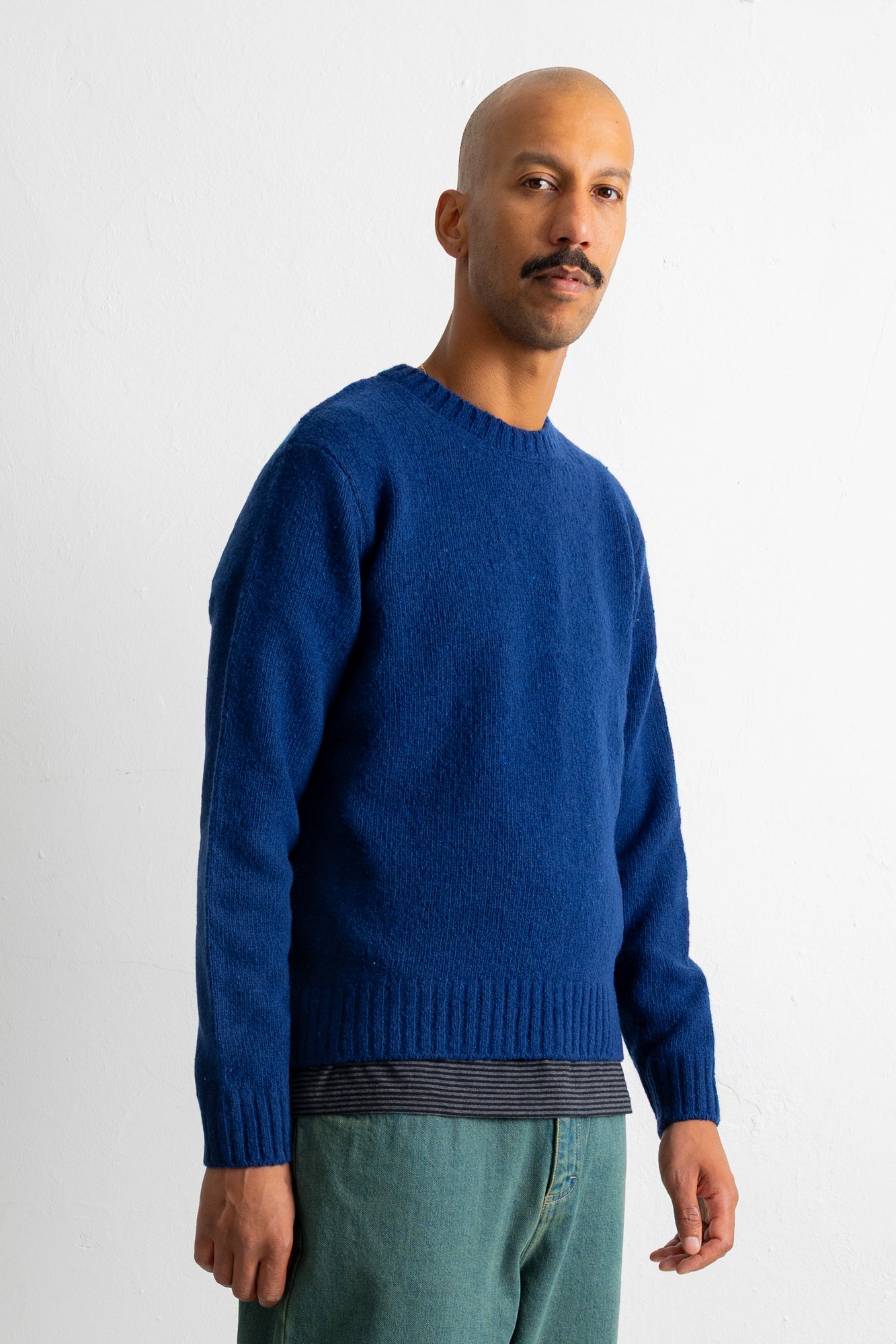 A man wearing a vibrant royal blue knit crewneck sweater, paired with green trousers and a striped undershirt, standing against a plain white studio background.