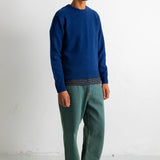 A man wearing a blue knit crewneck sweater, green trousers, and black canvas sneakers, standing against a plain white wall.