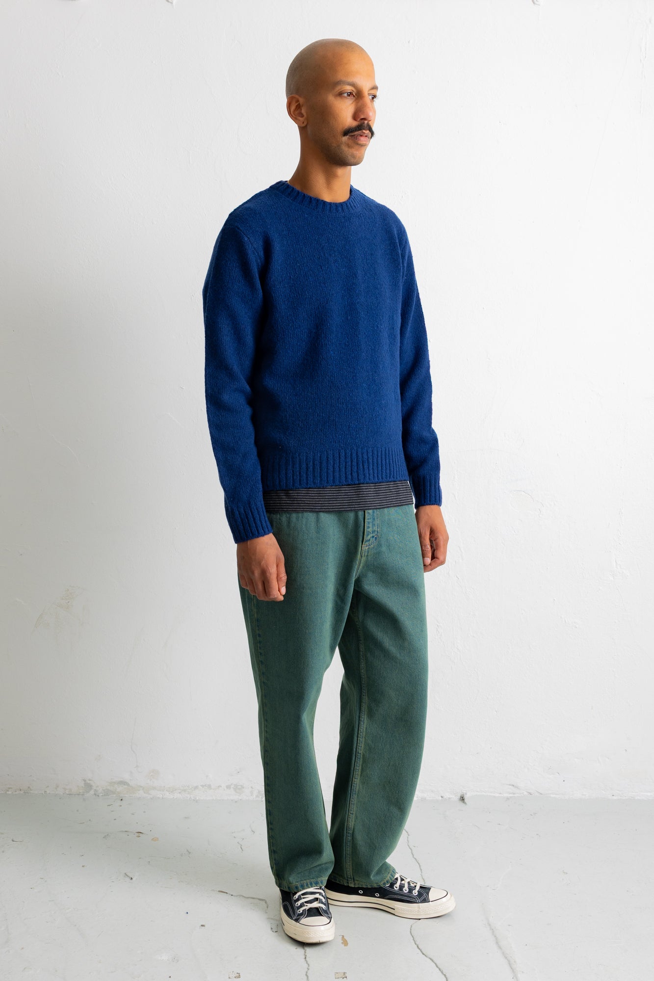 A man wearing a blue knit crewneck sweater, green trousers, and black canvas sneakers, standing against a plain white wall.