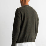 A man viewed from behind wearing a dark olive green knit sweater over a white t-shirt and blue jeans, against a plain white background.