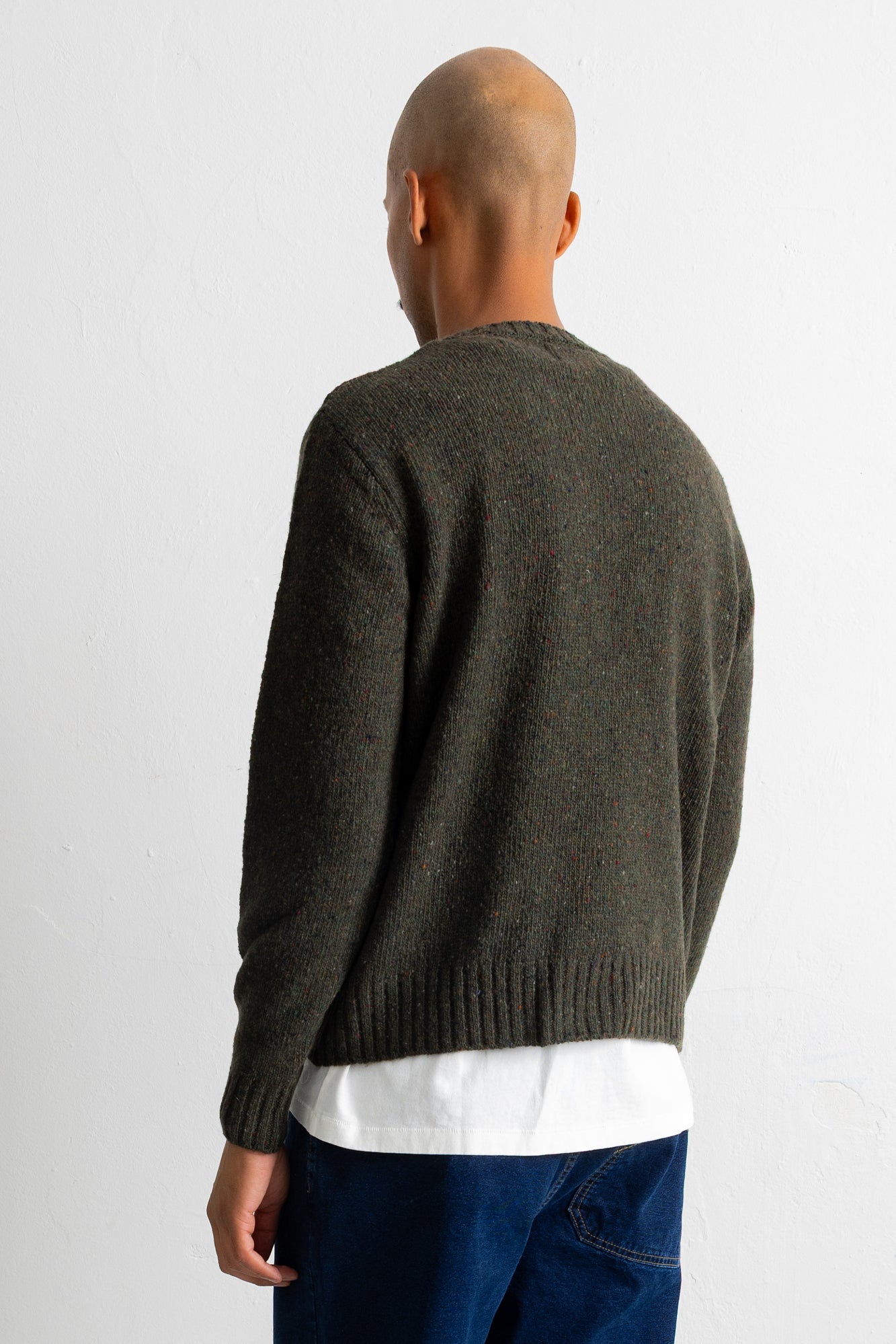 A man viewed from behind wearing a dark olive green knit sweater over a white t-shirt and blue jeans, against a plain white background.