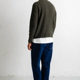 A man viewed from behind wearing a dark olive green knit sweater, a white t-shirt, dark blue jeans, and black canvas sneakers, standing against a plain white wall.