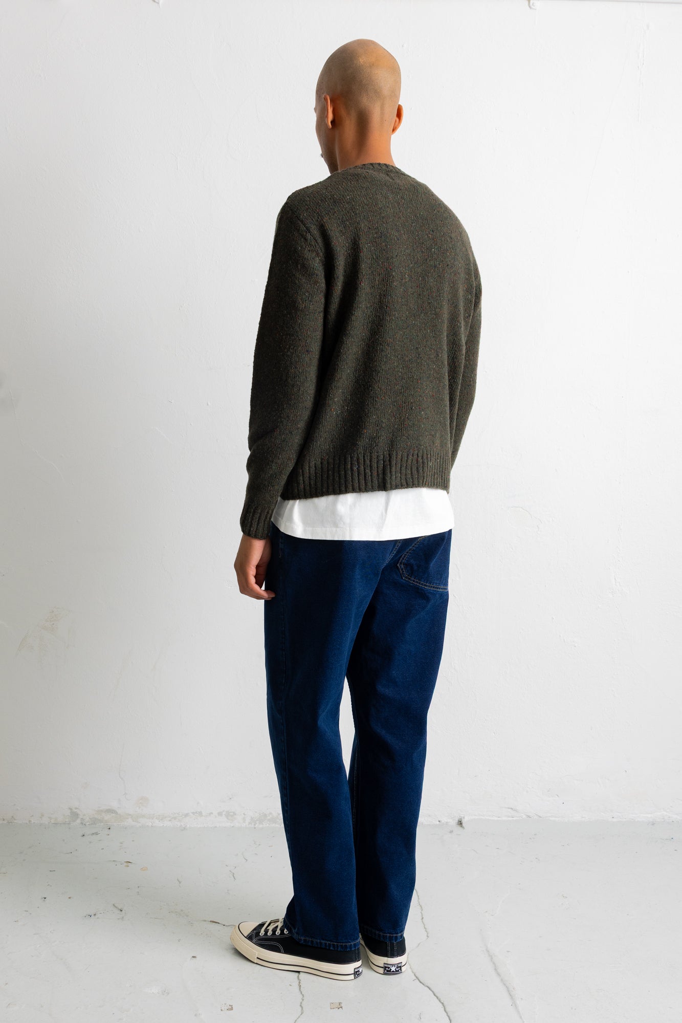 A man viewed from behind wearing a dark olive green knit sweater, a white t-shirt, dark blue jeans, and black canvas sneakers, standing against a plain white wall.