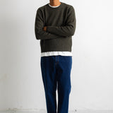 A man wearing a dark olive green knit crewneck sweater, blue jeans, and black and white sneakers, standing with arms crossed against a plain white studio wall.