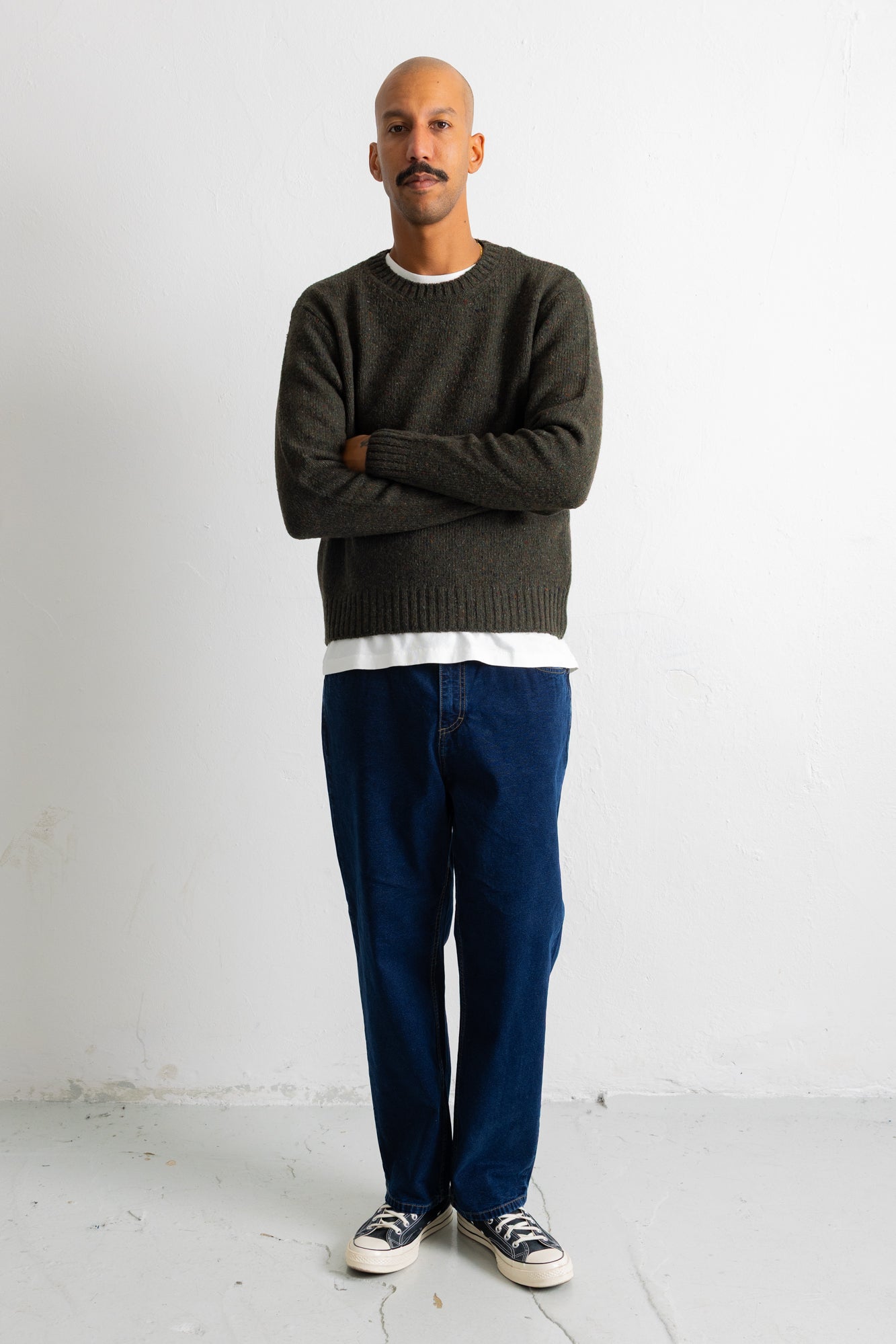 A man wearing a dark olive green knit crewneck sweater, blue jeans, and black and white sneakers, standing with arms crossed against a plain white studio wall.