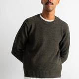 A man wearing a dark olive green crewneck sweater over a white t-shirt and blue jeans, standing against a plain light gray background.