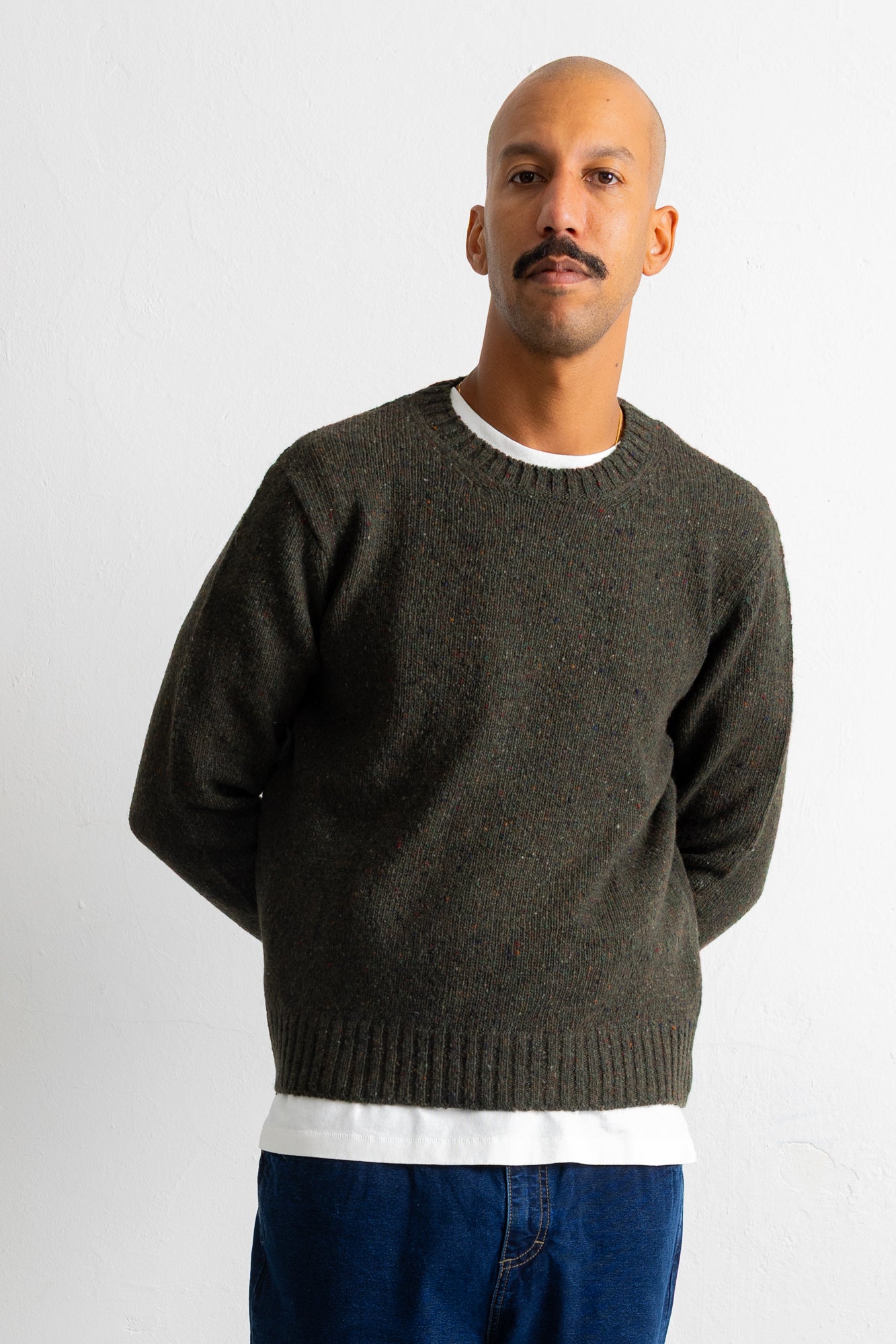 A man wearing a dark olive green crewneck sweater over a white t-shirt and blue jeans, standing against a plain light gray background.