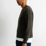A man in profile wearing a dark olive green knit sweater over a white shirt and blue jeans, standing against a plain light gray background.