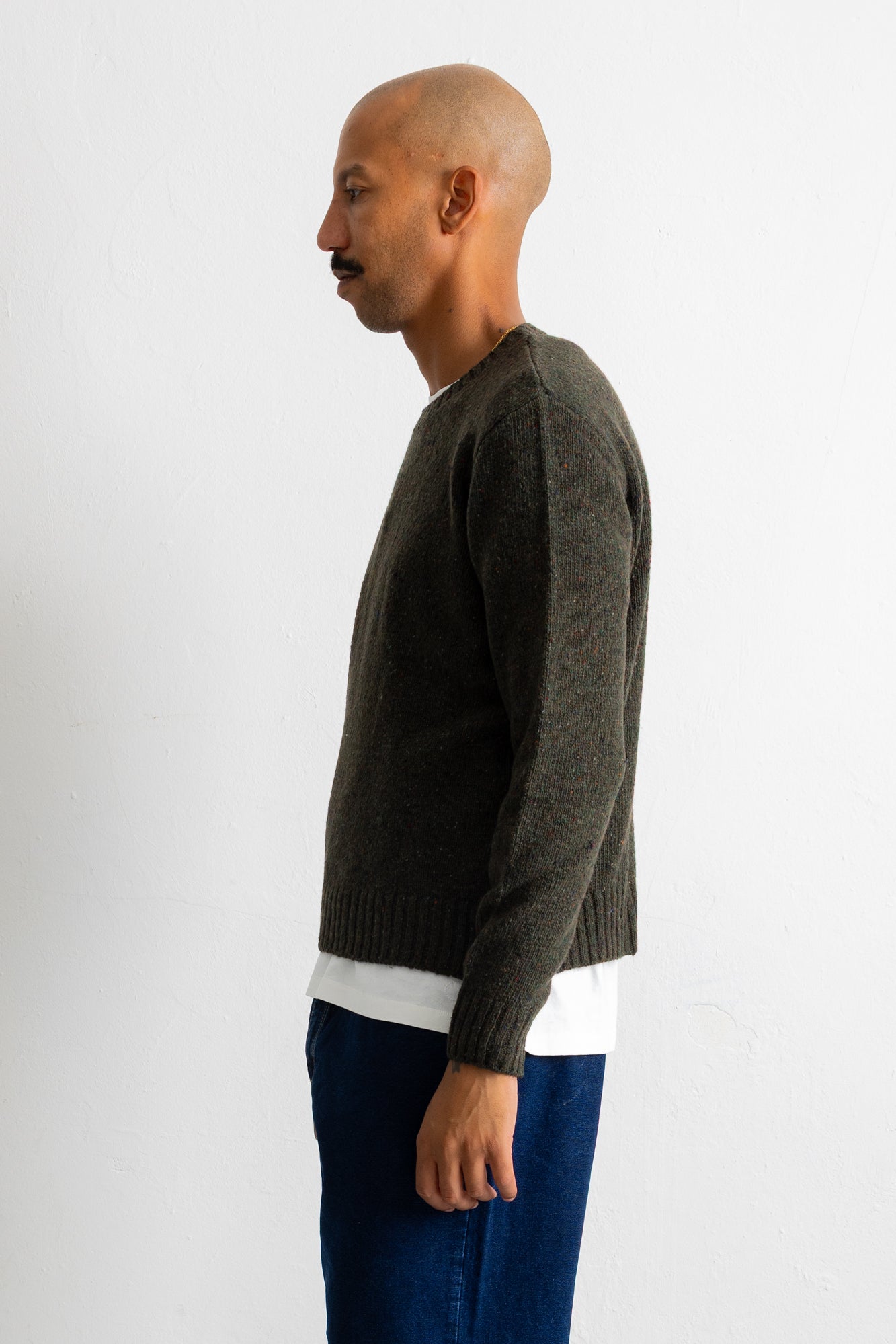 A man in profile wearing a dark olive green knit sweater over a white shirt and blue jeans, standing against a plain light gray background.