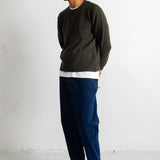 A man wearing a dark olive green knit crewneck sweater, a white undershirt, dark blue jeans, and black canvas sneakers, standing against a white wall.