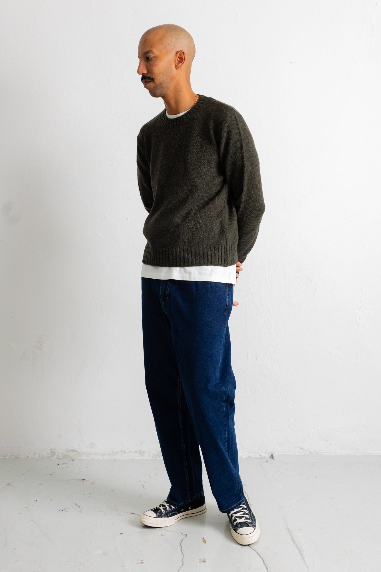A man wearing a dark olive green knit crewneck sweater, a white undershirt, dark blue jeans, and black canvas sneakers, standing against a white wall.