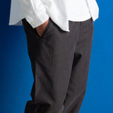 A person wearing dark charcoal gray trousers and a white textured shirt, standing against a solid blue studio background.