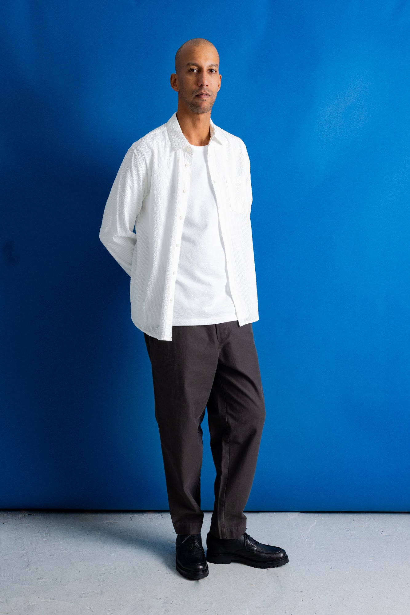 A man stands against a bright blue studio backdrop wearing a white long-sleeve button-up shirt over a white T-shirt, dark charcoal trousers, and black dress shoes.