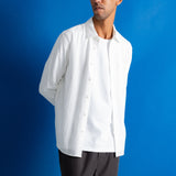 A man wearing a white long-sleeve button-up shirt and dark gray trousers, standing against a solid blue background.