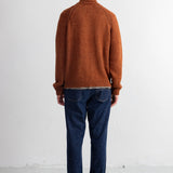 A man viewed from behind wearing a rust-orange knit sweater, dark blue jeans, and black shoes, standing against a plain white studio wall.